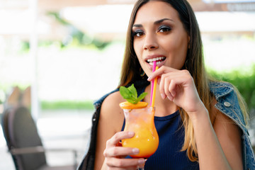 A beautiful young girl of European appearance is drinking an exotic summer cocktail while sitting at a table of a street summer cafe