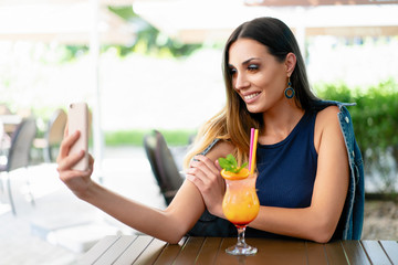 A beautiful smiling woman is drinking an exotic cocktail in an outdoor restaurant and is holding a mobile phone.