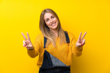 Woman with overalls over isolated yellow wall smiling and showing victory sign