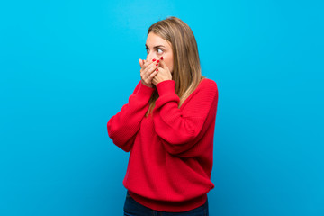 Woman with red sweater over blue wall covering mouth and looking to the side