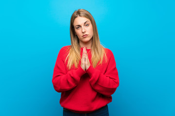 Woman with red sweater over blue wall keeps palm together. Person asks for something