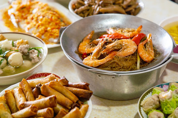 Casseroled prawns with Glass Noodels in Small Iron Pan with Fish, Fried Spring Rolls, Dim Sum, Seafood, Roasted Duck and Meatball Background on the Table.