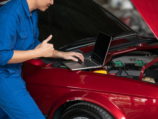 Close up of mechanic hand using laptop computer to check a car engine and showing thumbs up at the repair garage. Auto repair, car service and maintenance concept.