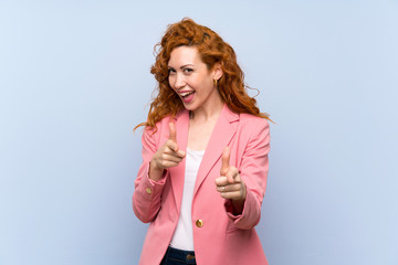 Redhead woman in suit over isolated blue wall pointing to the front and smiling