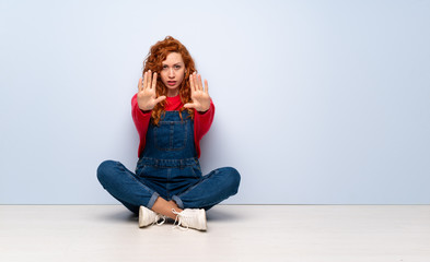 Redhead woman with overalls sitting on the floor making stop gesture and disappointed
