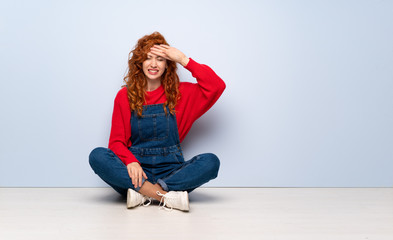 Redhead woman with overalls sitting on the floor with tired and sick expression