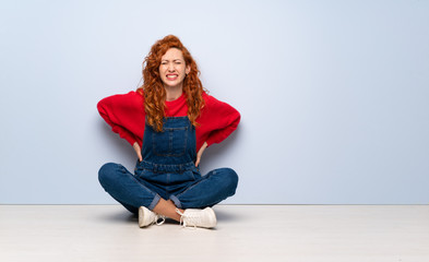 Redhead woman with overalls sitting on the floor suffering from backache for having made an effort