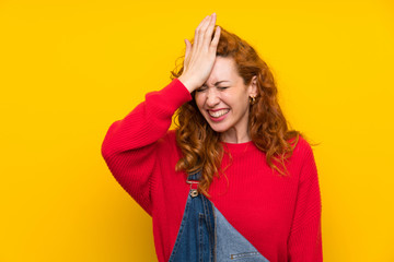 Redhead woman with overalls over isolated yellow wall having doubts with confuse face expression