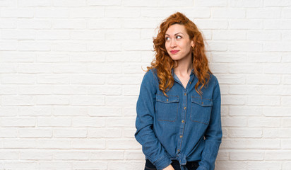 Redhead woman over white brick wall making doubts gesture looking side