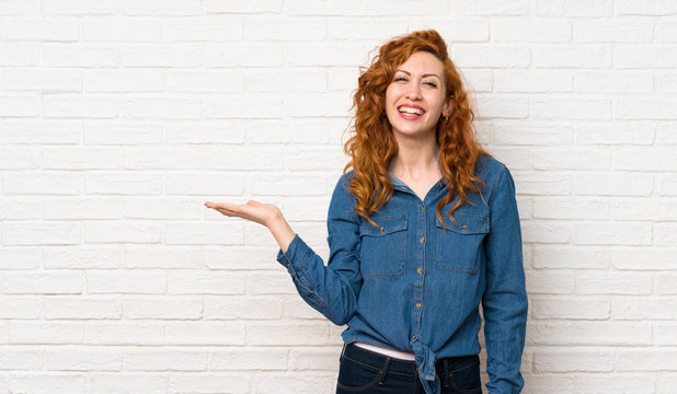 Redhead woman over white brick wall holding copyspace imaginary on the palm to insert an ad