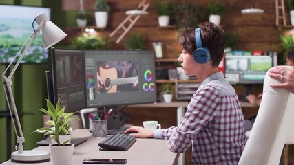 Female video editor and colorist puts her headphones on. In the background - professional post production house with a team working