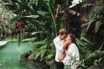 Happy couple kissing while relaxing in outdoor spa swimming pool surrounded with lush tropical greenery of Ubud, Bali. Luxury spa and wellness vacation retreat concept.