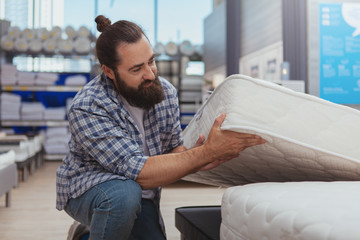 Household shopping concept. Mature bearded man examining orthopedic mattress at furnishings store....