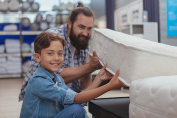 Adorable young cheerful boy smiling to the camera, examining orthopedic mattress for sale, shopping with his father for furniture. Father and son choosing new bed together