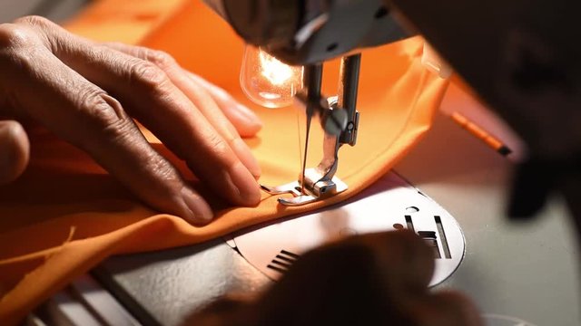 Woman sewing, Hands of old woman sewing dress from orange silk material on old vintage sewing machine close up