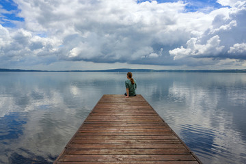 Naklejka premium A woman is sitting alone on a wooden walkway at the Lago di Bracciano in Italy