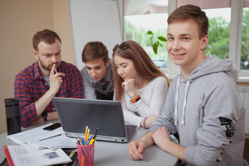 Cheerful teenage boy smiling to the camera, his classmates working on a project with teacher on the background. Happy teen male student enjoying studying at high school