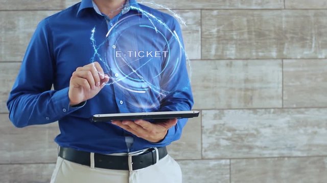 Man activates a conceptual hologram with text E-ticket. The guy in the blue shirt and light trousers with a holographic screen gadget on the background of the wall