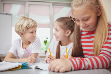 Gossip, sharing secrets concept. Two young schoolgirls whispering at art class lesson. Young schoolgirl drawing, while her friends chatting quietly on the background.