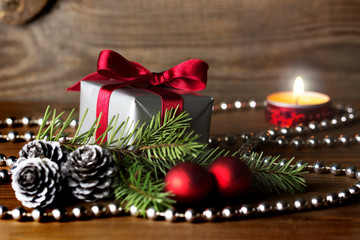 Christmas composition: a gift wrapped with red ribbon, silver cones, red baubles and a fir tree branch - burning candle in background