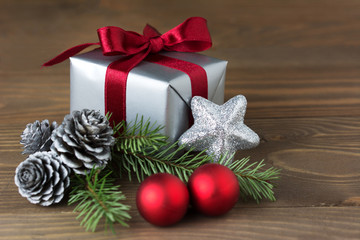 Christmas composition: a gift wrapped with red ribbon, silver cones, red baubles and a fir tree branch - isolated on wooden background