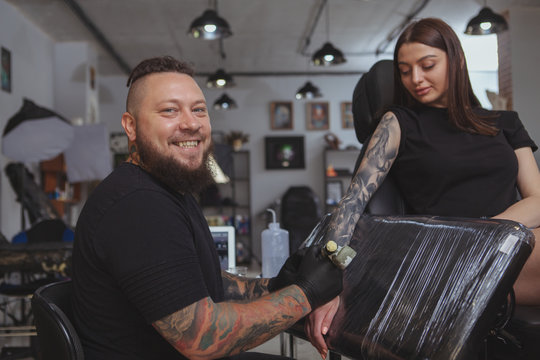 Cheerful bearded tattooed man smiling to the camera over his shoulder, tattooing young pretty woman at his salon. Professional tattoo artist enjoying doing tattoos at his workshop