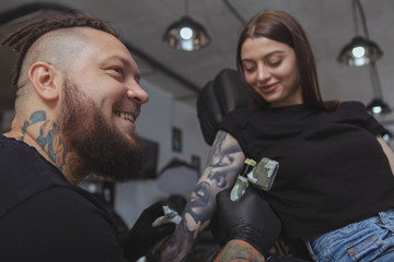 Fototapeta na wymiar Close up of a cheerful bearded tattoo artist enjoying working at his tattoo salon, working with female client. Professional tattooer tattooing attractive woman