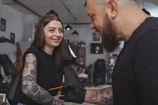 Attractive happy young woman laughing, talking to her tattoo artist showing him healed artworks tattooed on her arm. Rear view shot of a bearded tattooer examining healed tattoos on the arm of female 