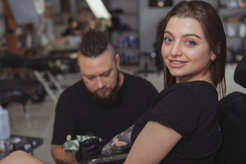 Close up of a happy beautiful woman smiling to the camera while getting arm sleeve tattoo done by professional tattoer. Cheerful female client being tattooed by tattooer