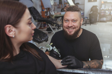 Happy bearded male tattoo artist laughing, talking to his female customer while tattooing her arm. Professional tattooer enjoying working at his ink salon