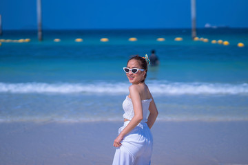 Summer photo of stylish pretty woman in sunglasses, wearing white dress, enjoying the summer days, on the sand beach, near the sea