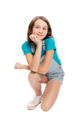 Smiling teen girl in denim overalls squats. Isolated on a white background. Vertical.