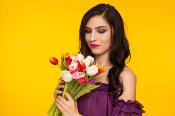 Beautiful brunette woman holding a bouquet of tulips in her hands on a yellow background. Female dressed in a purple, thin dress. March 8, the day matri.