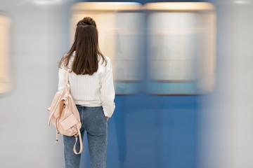 young white girl with long hair. With a backpack. Standing at the metro station and waiting for the train. The train is out of focus to convey the effect of the reality of the movement.