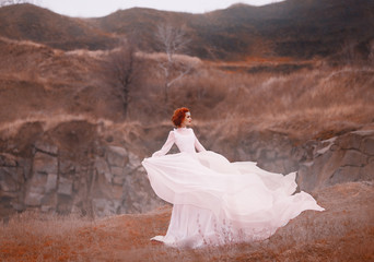 Red-haired beauty in a white vintage dress, stands on top of a hill, Background of the rocky mountains. Luxurious outfit with a long train flutters in the wind. Light flying silk fabrics. warm colors