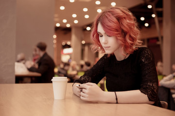 Beautiful young girl, female student drinking hot coffee, drink sitting on food court in a shopping center. Looks at the smartphone and correspond with the social network. Red, orange hair.