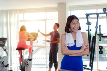 young happy smiling asian woman slim body with towel crossing arms in fitness gym with people on machine bicycle background, bodybuilder, healthy lifestyle, exercise, workout, sport training concept