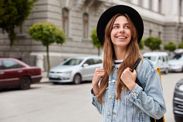 Outdoor shot of beautiful European woman strolls through city, spends free time, recreats during vacation, wears hat and denim jacket, walks around cozy street, blurred background, transport.