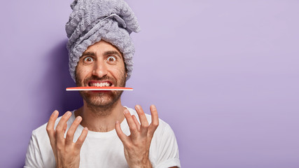Photo of funny unshaven guy keeps nail file in mouth, annoyed with beauty procedures, going to do manicure, wears bath towel on head, visits beauty salon, isolated on purple wall with empty space
