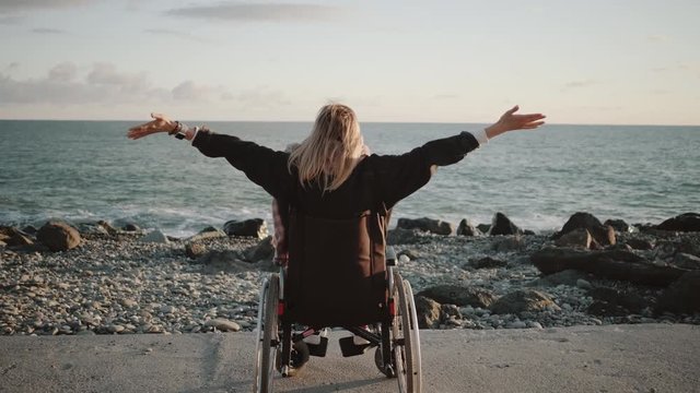 Alone disabled woman is sitting in wheelchair and holding hands up on sea coast