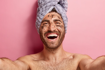 Healthy positive man makes selfie, applies coffee scrub on face skin, has cleansing procedures, poses topless against pink background with towel on head. Cosmetology, masculinity, beauty concept