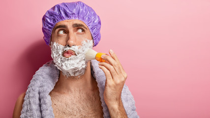 Close up shot of handsome man applies shaving gel on face with brush, wears shower cap, focused aside, has towel around neck, enjoys morning routine, protects skin from irritation, isolated on pink