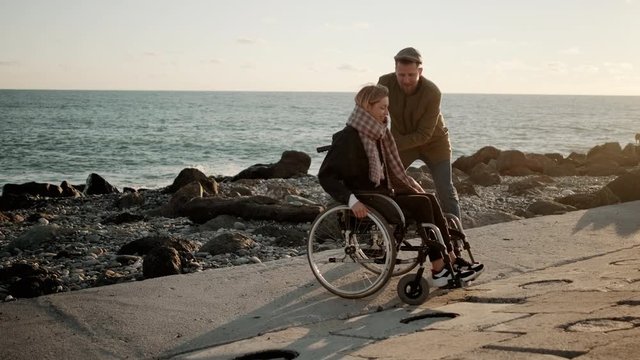 Loving man is whirling wheelchair with young woman on sea coast, rejoicing