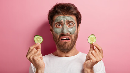 Upset man has clay mask on face, holds two cucumber slices, visits spa salon, experiments new...