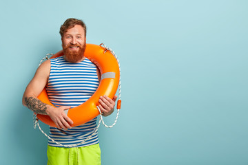 Positive bearded red haired guy wears striped sailor vest, carries orange lifebuoy, stands over...