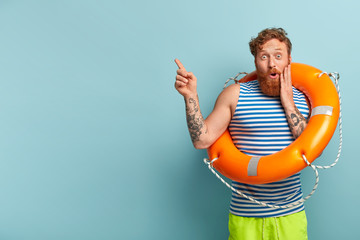 Safe sinking concept. Astonished red haired man holds orange lifebuoy, maintains safety, wears...