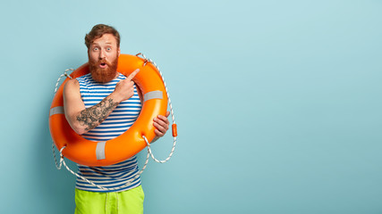 Surprised red haired man points aside away, poses with safety equipment, points aside on blank...