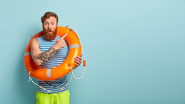 Surprised red haired man points aside away, poses with safety equipment, points aside on blank space, has tattoo on arm, shows direction somewhere, has astonished facial expression. Beach vacation