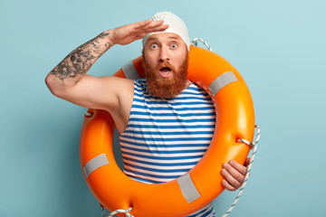 Shocked bearded red haired man looks surprisingly into distance, keeps palm near forehead, mouth wide opened, wears striped casual t shirt, poses with lifeguard, isolated over blue background.