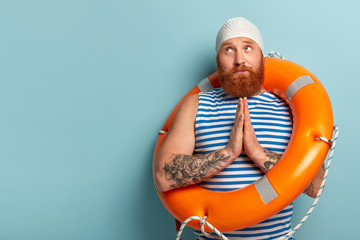 Hopeful handsome male swimmer keeps palms pressed together, prays for safe swimming, recreats in resort place, wears sailor vest, white swimcap, carries inflated lifebuoy, blank space for your promo - obrazy, fototapety, plakaty